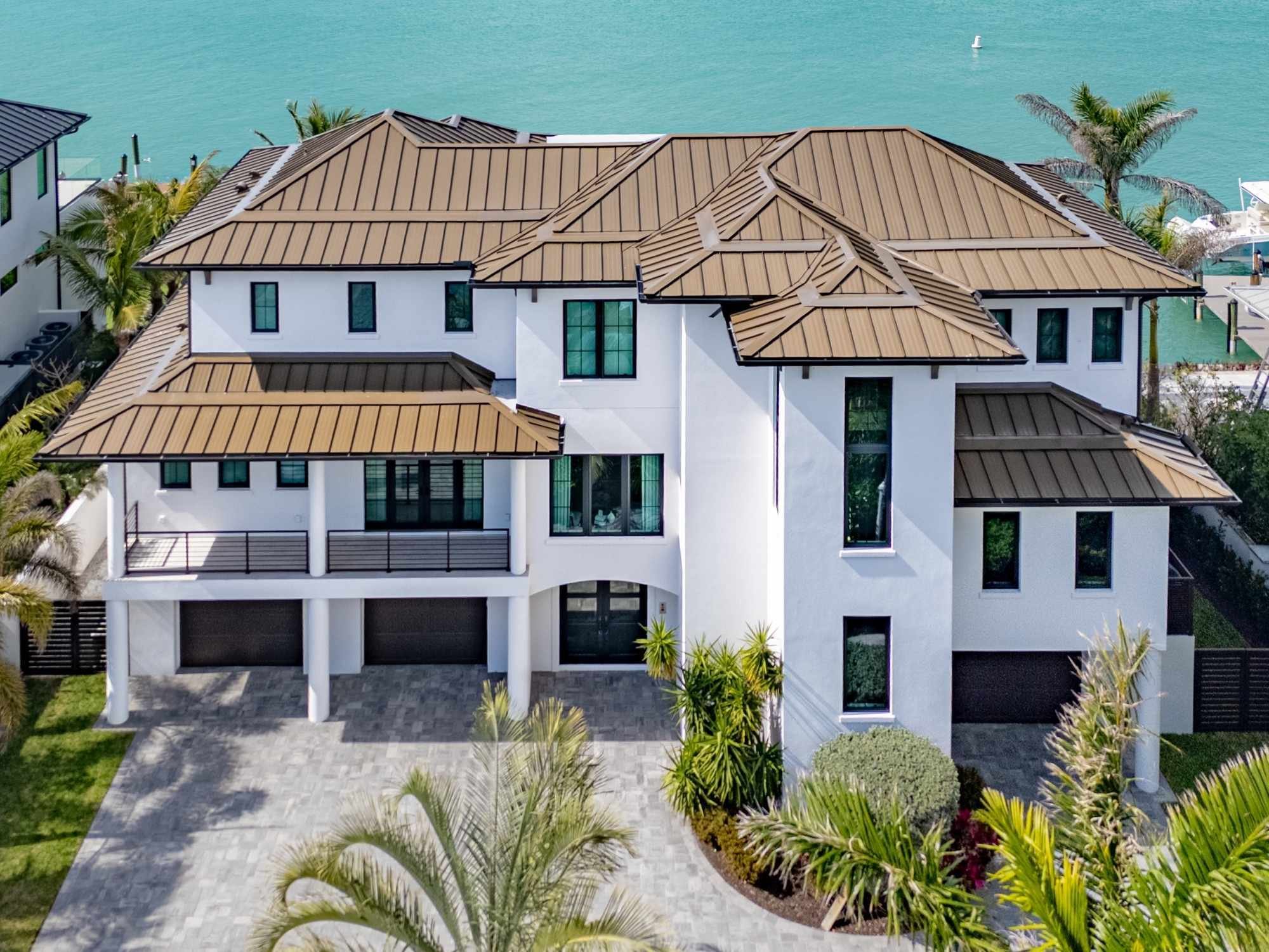 Waterfront custom home with white stucco, bronze metal roofing, multiple garages, and tropical landscaping in Southwest Florida.